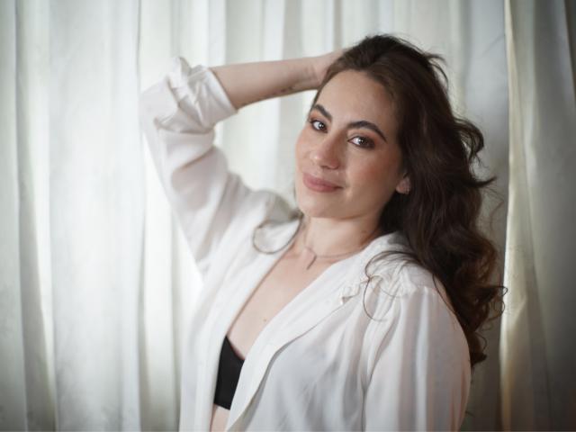 LuciaPerth with wavy brown hair, wearing a white shirt over black bra, hand in hair, subtle smile, soft light, natural makeup, casual, confident, sexy, slightly open shirt.