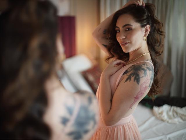 LuciaPerth with wavy brown hair, pink dress, and tattoos on her arm and shoulder, smiling at herself in a mirror, holding her hair up, in a softly lit room.