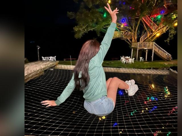 AnyaJay with long brown hair, green hoodie, denim shorts, white sneakers, sitting on black grid, raising arm, colorful lights on grid, tree and wooden structure in background.