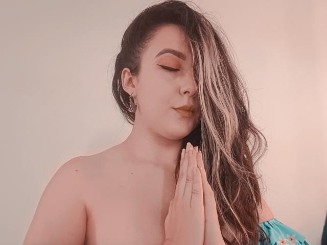 MabyLegrand with long brown hair, eyes closed, and hands in prayer, topless, showing tits, wearing gold earrings, and a blue floral garment on her shoulder. Soft pink lighting.