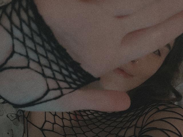 FionaLaNice mit heller Haut, die schwarze Netzstrümpfe und Dessous trägt, ihre Brüste sind sichtbar, sie hat einen Nasenring und dunkles Haar, in einer schwach beleuchteten, intimen Umgebung..