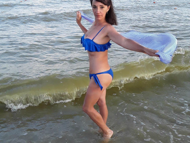 MissieLorrie, en maillot de bain bleu, tenant un objet gonflable blanc, se tient dans une eau peu profonde avec de petites vagues, montrant ses seins et ses fesses, et regarde l'appareil photo avec un léger sourire..