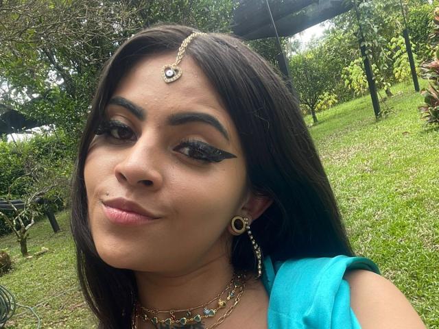 CoriPaige has dark brown hair, thick eyebrows, black winged eyeliner, and wears a gold forehead jewel. She's in a blue top, grassy background, and has a subtle smile.