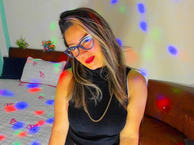Contelaya with blue glasses, red lipstick, black sleeveless top, and gold chain, sitting on brown couch, colorful laser lights on her face and bed.