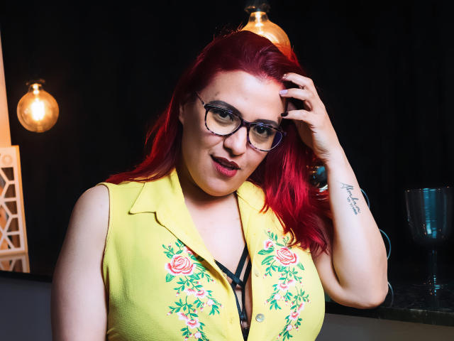 SammyOwens with red hair, glasses, and a yellow floral top, touching her head, showing a tattoo on her right wrist, smiling with red lipstick. Boobs, no pussy visible.