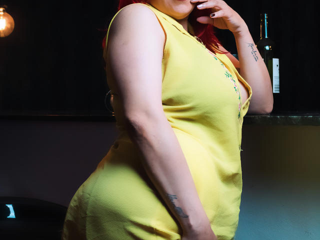 SammyOwens in a yellow dress, showing her ass and tits, with red hair, a tattoo on her wrist, and a hand near her mouth, standing in a dimly lit room.