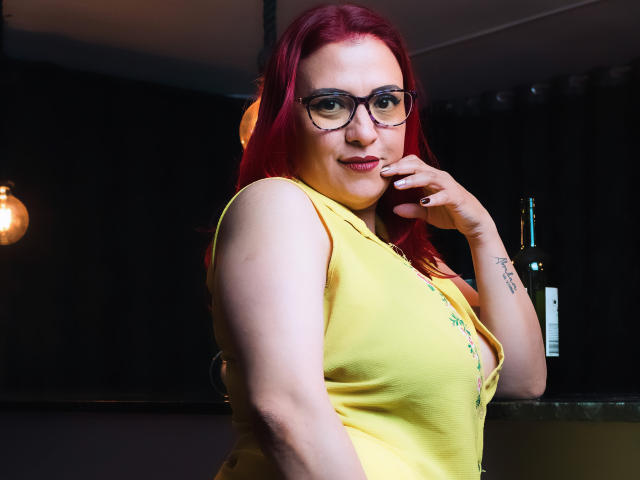 SammyOwens with red hair, glasses, yellow top, hand near mouth, small tattoo on right wrist, bottle in background, light bulb on left, slight smile, pale skin, curvy.