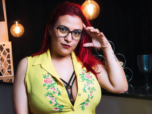 SammyOwens with red hair, glasses, yellow shirt with rose embroidery, black strappy bra, hand in hair, cute, large tits, fair skin, black background, hanging lights, subtle tattoo.