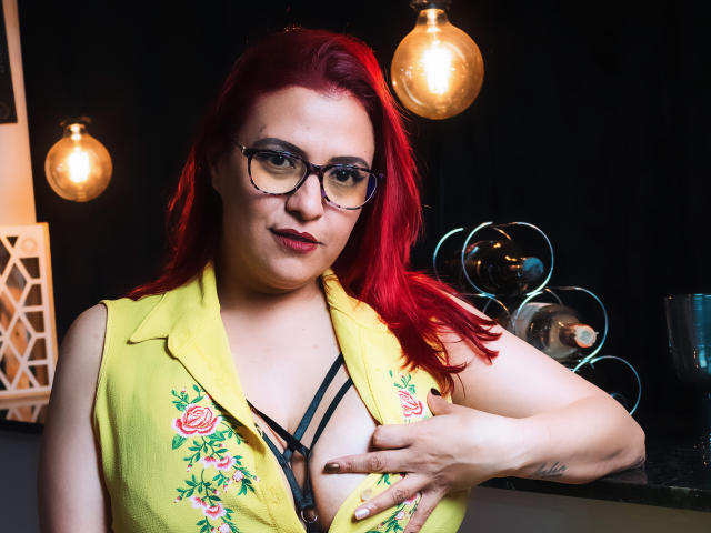 SammyOwens with red hair, glasses, and a yellow vest exposing black strappy lingerie, holding her tits with a hand, a tattoo on her right wrist, and a smug expression.