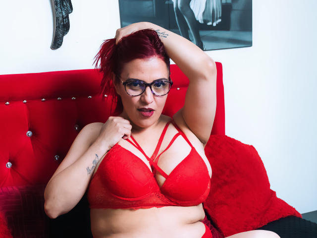 Sammy Owens, com cabelo ruivo, óculos e um sutiã de renda vermelho, segurando o cabelo para cima, mostrando os seios, sentada em uma cama vermelha com estofamento, olhando para a câmera..