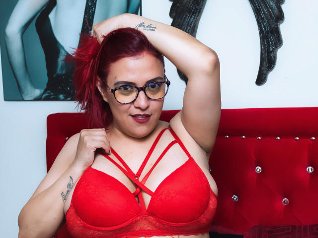 SammyOwens with red hair, glasses, and red lace bra, showing off her big tits, tattoo on right arm, hand in hair, sitting on red tufted couch.