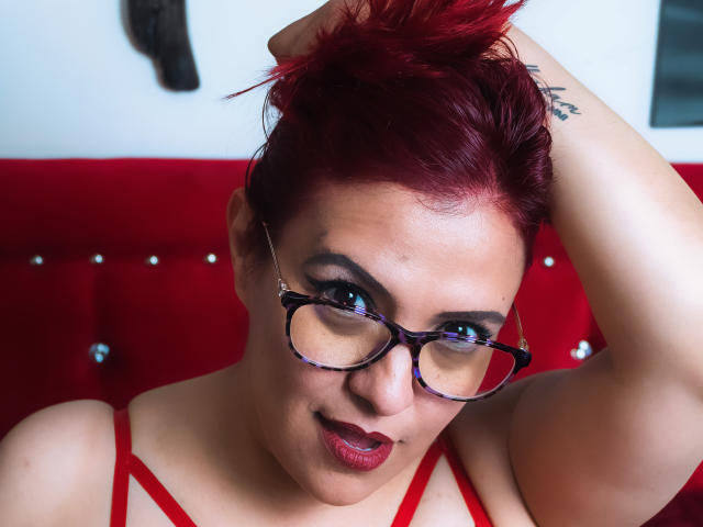 SammyOwens with red hair, glasses, and red lipstick, raising an arm to her head, showing a tattoo on her arm, wearing a red strappy top, looking at the camera.