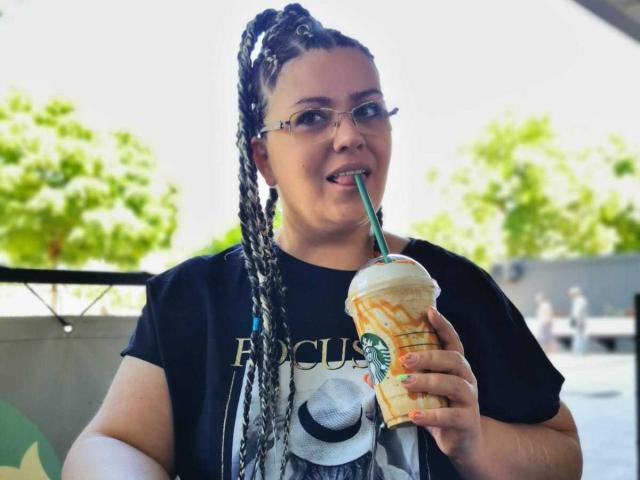 Carolina69 with long braids, sipping iced coffee through a green straw, tongue out, wearing glasses and a black t-shirt with a hat graphic, outdoors.