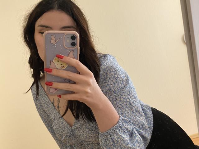 Rubbi with long dark hair, blue floral top, red nails, holding phone with cat case, showing tits, mirror selfie, black skirt, white wall, wooden floor, looking at phone.