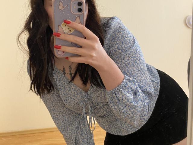 Una donna con capelli scuri ondulati, una camicetta floreale blu e una gonna nera, è china in avanti, mostrando il seno, tiene in mano un telefono con le unghie rosse e una custodia per telefono carina..