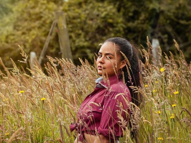 ArielleWilde with long black hair in a ponytail, wearing a maroon shirt, standing in a tall grass field, her tits slightly visible, looking to the side.
