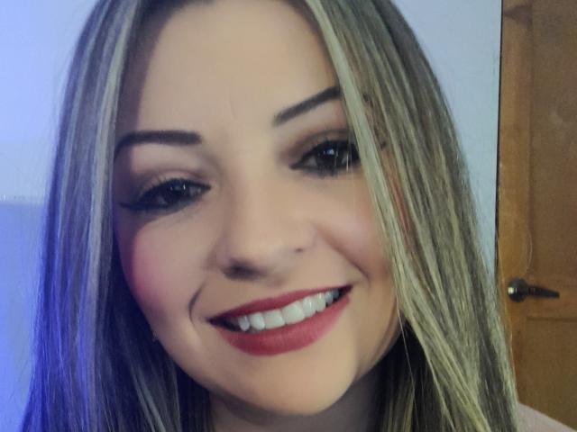 NatalyHami hat blondes Haar, blaue Augen und ein süßes Lächeln mit rotem Lippenstift. Sie zeigt ihre weißen Zähne und hat einen kleinen Schönheitsfleck in der Nähe ihres rechten Auges..
