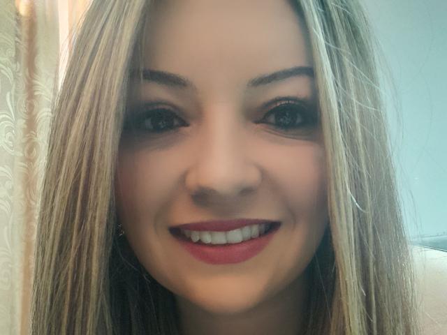 NatalyHami tem cabelo loiro, olhos castanhos e batom vermelho, sorrindo com os dentes à mostra. Seu nariz é centralizado e ela está usando uma maquiagem discreta..
