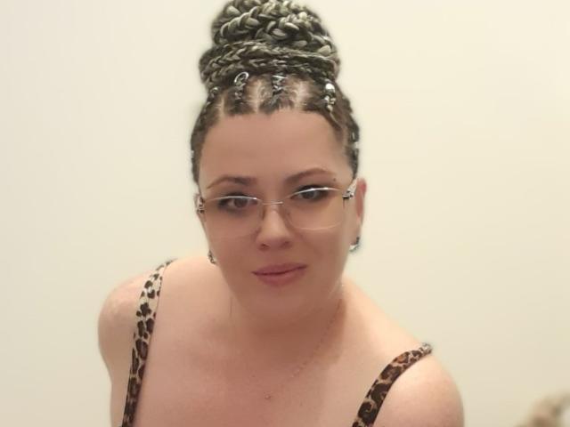 Carolina69 has braided-updo, wearing glasses, leopard-print top, showing her tits, fair skin, pink lipstick, and subtle smile, against plain background.