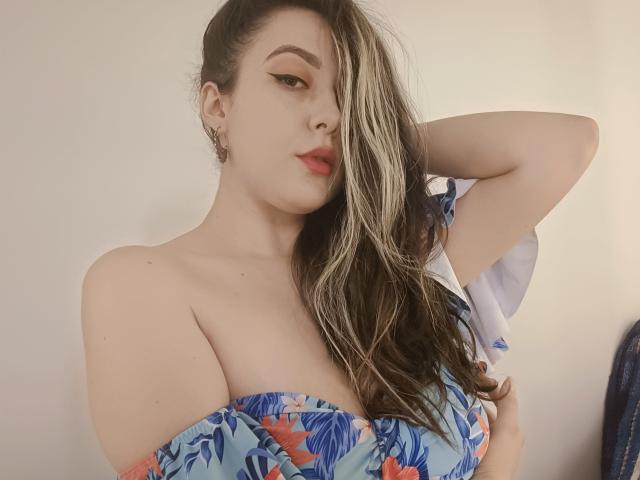 MabyLegrand with wet, blonde-highlighted hair, blue floral off-shoulder top, arm raised, showing cleavage, full red lips, eyeliner, and earrings, in a softly lit, neutral background.