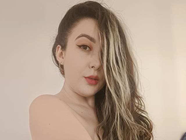 MabyLegrand ha lunghi capelli biondi e castani, ondulati, che coprono un occhio, un audace eyeliner arancione e labbra rosse, con le spalle scoperte e un'espressione neutra. Una luce soffusa illumina il suo viso..