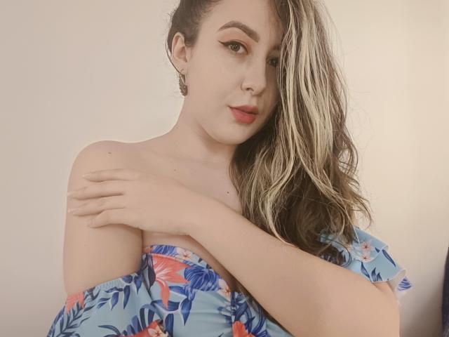 MabyLegrand with blonde and brown wavy hair, wearing a blue floral off-shoulder top, arms crossed, showing tits, red lipstick, eyeliner, and earrings, against a plain background.