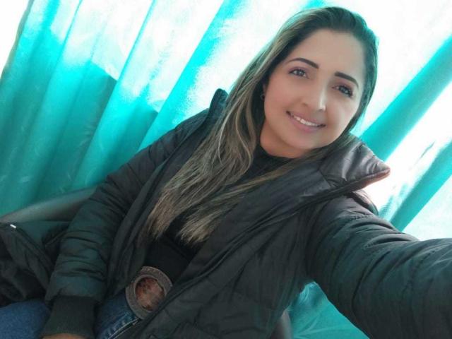 StefanyCaruso ha lunghi capelli castani, indossa una giacca nera e una maglietta nera, sta sorridendo e si sta scattando un selfie, mostrando il suo viso carino e i suoi piccoli seni, con delle tende color verde acqua sullo sfondo..
