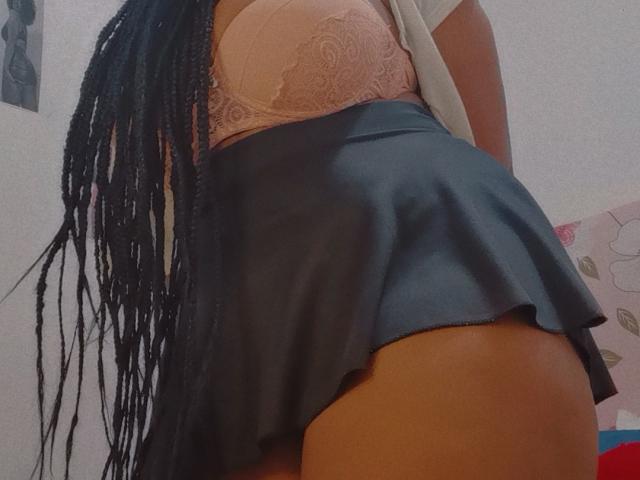 Eilin's pink lace bra and blue skirt show her curvy ass and thick thighs, with long black braids falling over her side. Her tan skin is visible.