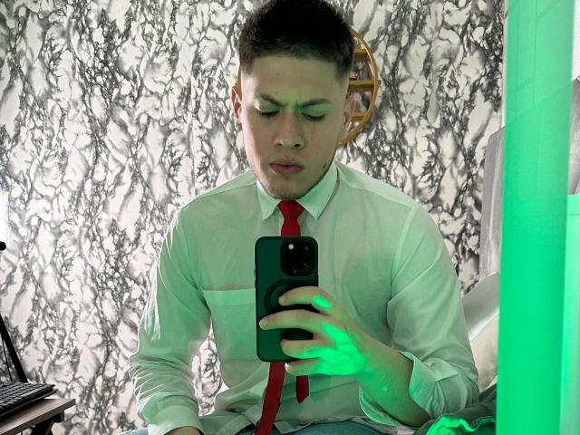 SebastiianMaxime, com cabelo curto e escuro, camisa branca, gravata vermelha e expressão séria, segurando um telefone preto, com uma luz verde na mão, e uma parede de fundo com padrão de mármore..