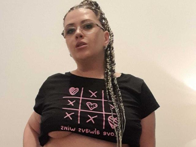 Carolina69 with braided hair, glasses, and a black crop top showing pink symbols, has small tits and a slightly pouty expression, standing against a plain wall.