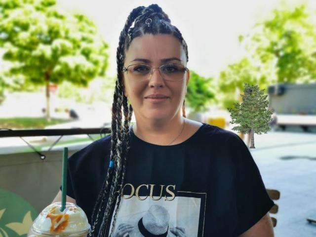 Carolina69 with long braided black and white hair, wearing glasses and a black t-shirt, holding a drink with a green straw, smiling outside.