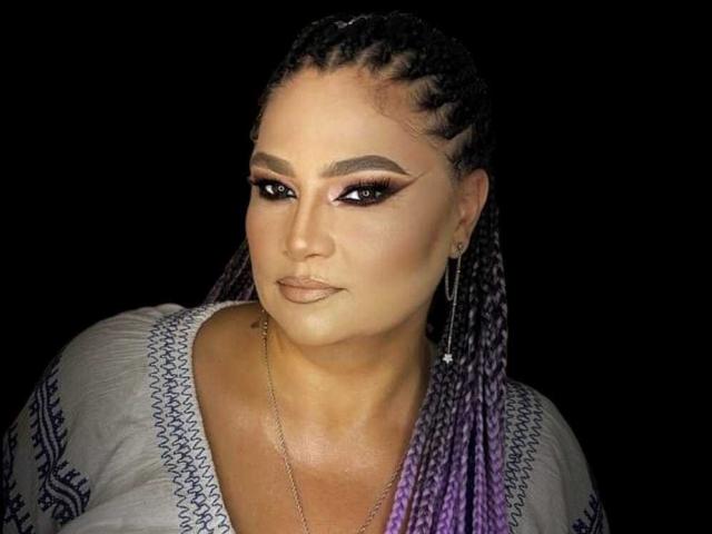 HellenBooty69 has braided purple and black hair, thick eyebrows, bold eyeliner, and purple eyeshadow. She's wearing a white top with blue patterns and a silver necklace.