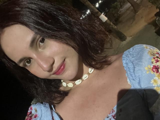EstefaniaRamosHot tiene el pelo oscuro y ondulado, piel clara, lápiz labial rojo, un vestido con estampado floral, mangas azules y un collar de conchas. Está sonriendo con una expresión linda y sensual..