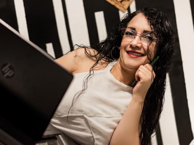 TitaNaughty with wet black hair, glasses, red lipstick, white sleeveless top, smiling at a laptop, holding a green pen in her right hand, with a confident, cute expression.