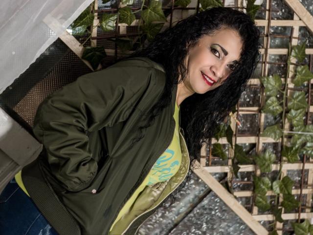 TitaNaughty with curly black hair, green jacket, yellow shirt, and blue jeans, smiling at camera, hands in pockets, red lipstick, green eyes, standing in front of wooden lattice.
