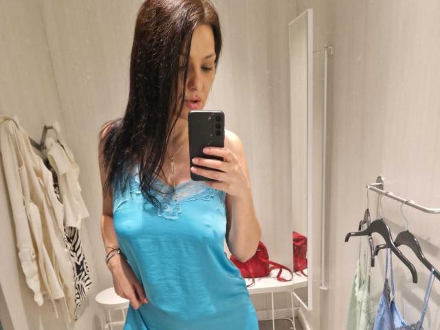 JoliDanielle with wet brown hair, blue dress, showing her tits in a mirror selfie, holding a black phone, red bra on a white table, clothes hanging on right.