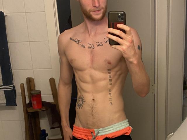 KillianNolan, ohne Hemd, hält ein Telefon, Spiegel-Selfie, muskulös, Tattoos auf Brust und Seite, orangefarbene Hose, rote Getränkedose im Hintergrund, leicht sichtbarer Holenstuhl, weiße Fliesenwand..