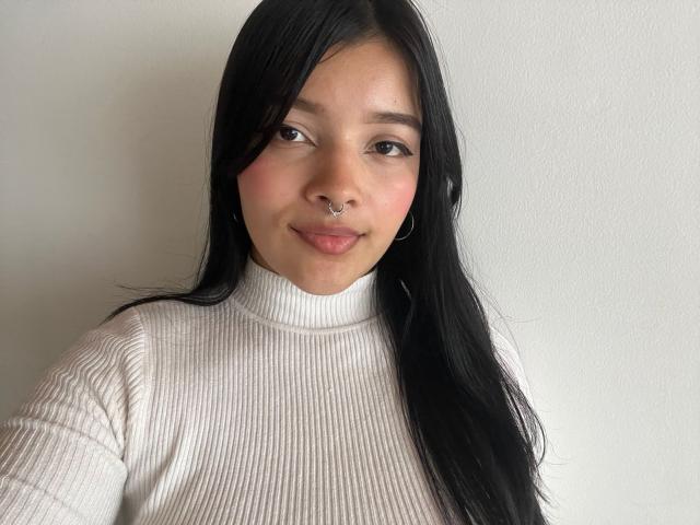 Emma, con cabello negro, un piercing en la nariz y aros, lleva un cuello alto blanco texturizado y sonríe suavemente. Sus senos no son visibles. Tiene un rostro lindo y atractivo..