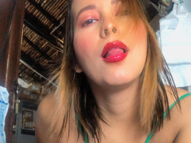 SaritaVelez with tongue out, red lipstick, nose ring, green strap top, blonde hair, slightly tilted head, looking up, soft light, slightly flushed skin, medium breast size, casual selfie.