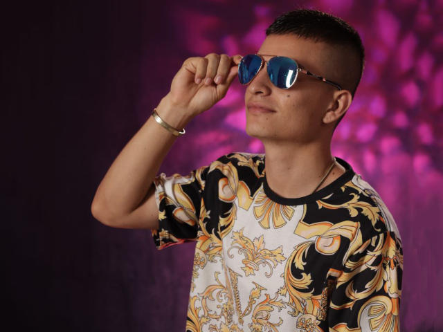 ShatSex mit kurzen, braunen Haaren, der reflektierende, blaue Sonnenbrillen, ein buntes, blumiges Hemd und ein silbernes Armband trägt und mit einem selbstbewussten, leicht arroganten Ausdruck seine Brille zurechtrückt..
