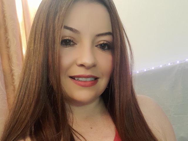 NatalyHami has long brown hair, light skin, and a cute smile with pink lipstick. She's wearing a red top and has subtle eyeliner and mascara.