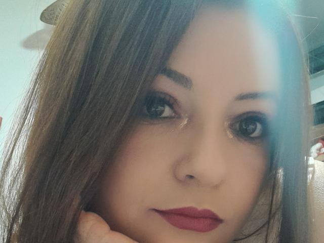 NatalyHamis Gesicht mit braunen Haaren, blauen Augen, dezentem Make-up, rotem Lippenstift und einer Hand unter dem Kinn, die leicht zur Seite blickt, mit weichem Licht im oberen rechten Bereich..