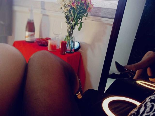 Las piernas negras y los tacones altos negros de Evangeline Cors se reflejan en un espejo, con una mesa roja, velas y un ramo de flores en el fondo. Su trasero está en primer plano..