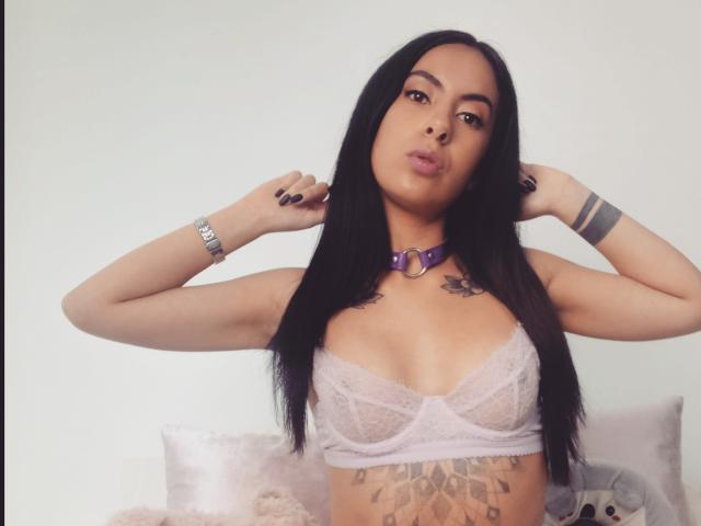 Sarah Luna mit langen, schwarzen Haaren, voller Lippen, die einen violetten Choker trägt, ein Spitzen-BH, der ihre Brüste zeigt, ein Tattoo auf der Brust, ein Tattoo am Arm und eine Armbanduhr..