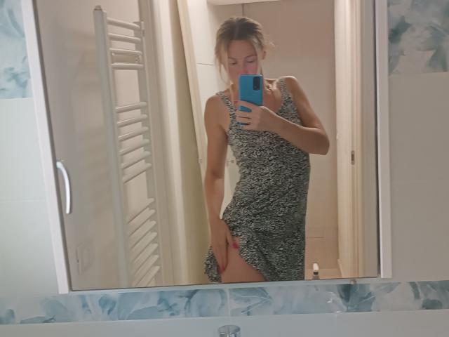 Saveta, vestindo um vestido verde e preto com estampas, segurando um telefone azul, levantando a saia para mostrar sua coxa nua e roupa íntima rosa. Selfie no espelho em um banheiro..