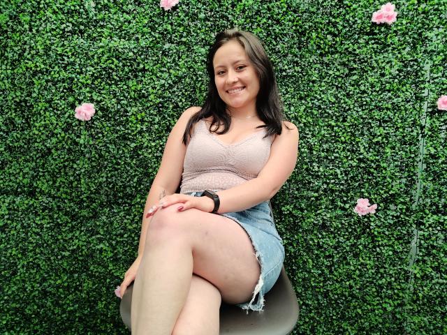 AnhaliaSkarleth, com seios médios, vestindo uma blusa de renda rosa e uma saia jeans azul, sentada em um banquinho, sorrindo, em frente a uma parede verde com flores rosa..