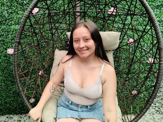 AnhaliaSkarleth, com cabelos castanhos longos e pele clara, está sentada em uma cadeira de balanço de vime, sorrindo, vestindo uma blusa rosa com detalhes em renda e shorts jeans, mostrando seios pequenos e tatuagens..