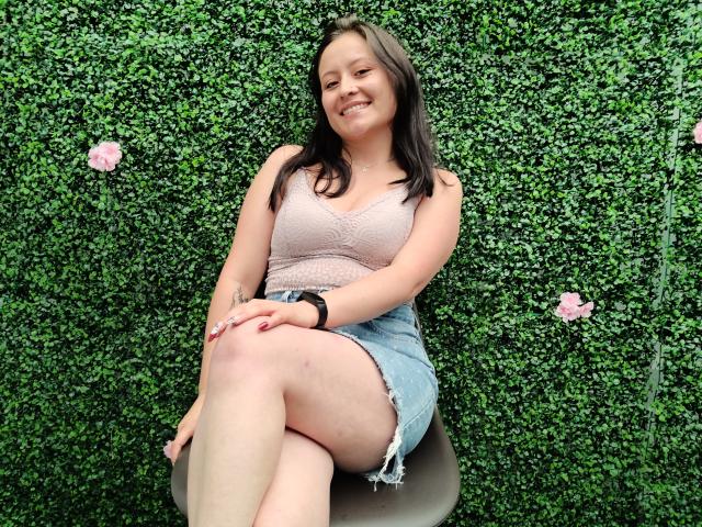 AnhaliaSkarleth, con un seno di media grandezza, indossa una maglia di pizzo rosa e una gonna di jeans blu, è seduta su una sedia, sorride, con le gambe incrociate, un braccialetto nero, una parete verde con fiori rosa..