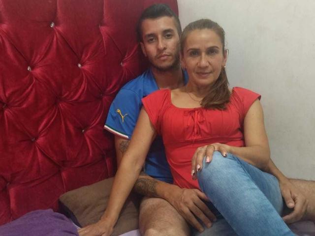 Tifany und Stiven sitzen auf einem Bett mit einem roten, gepolsterten Kopfteil. Stiven trägt ein blaues Hemd, Tifany ein rotes Oberteil und blaue Jeans. Seine Hand liegt auf ihrem Oberschenkel, ihre Hand auf seinem..