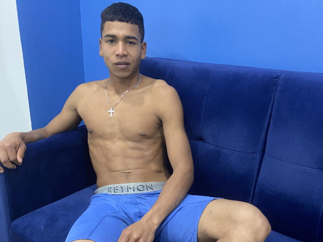 JhonBennet, shirtless, with a cross necklace, blue shorts, and a toned, slim, dark-skinned body, sitting on a blue couch, looking at the camera with a serious expression.
